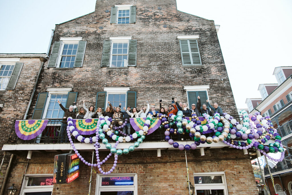 A group of guests in elaborate Mardi Gras costumes and wigs posing on a private balcony overlooking Bourbon Street, adorned with purple and green balloons.